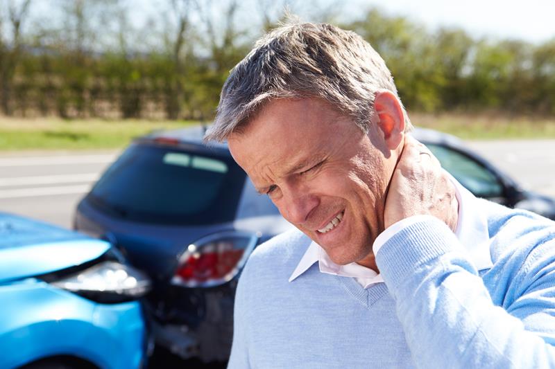 A man holds the back of his neck in pain, with two damaged cars visible in the blurred background, suggesting a car accident.