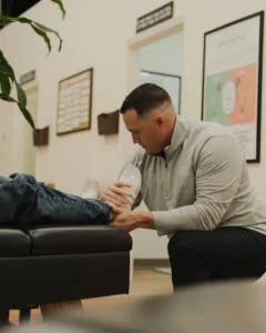 A male practitioner kneels, holding and examining a patient's foot on a black examination table in a clinic.