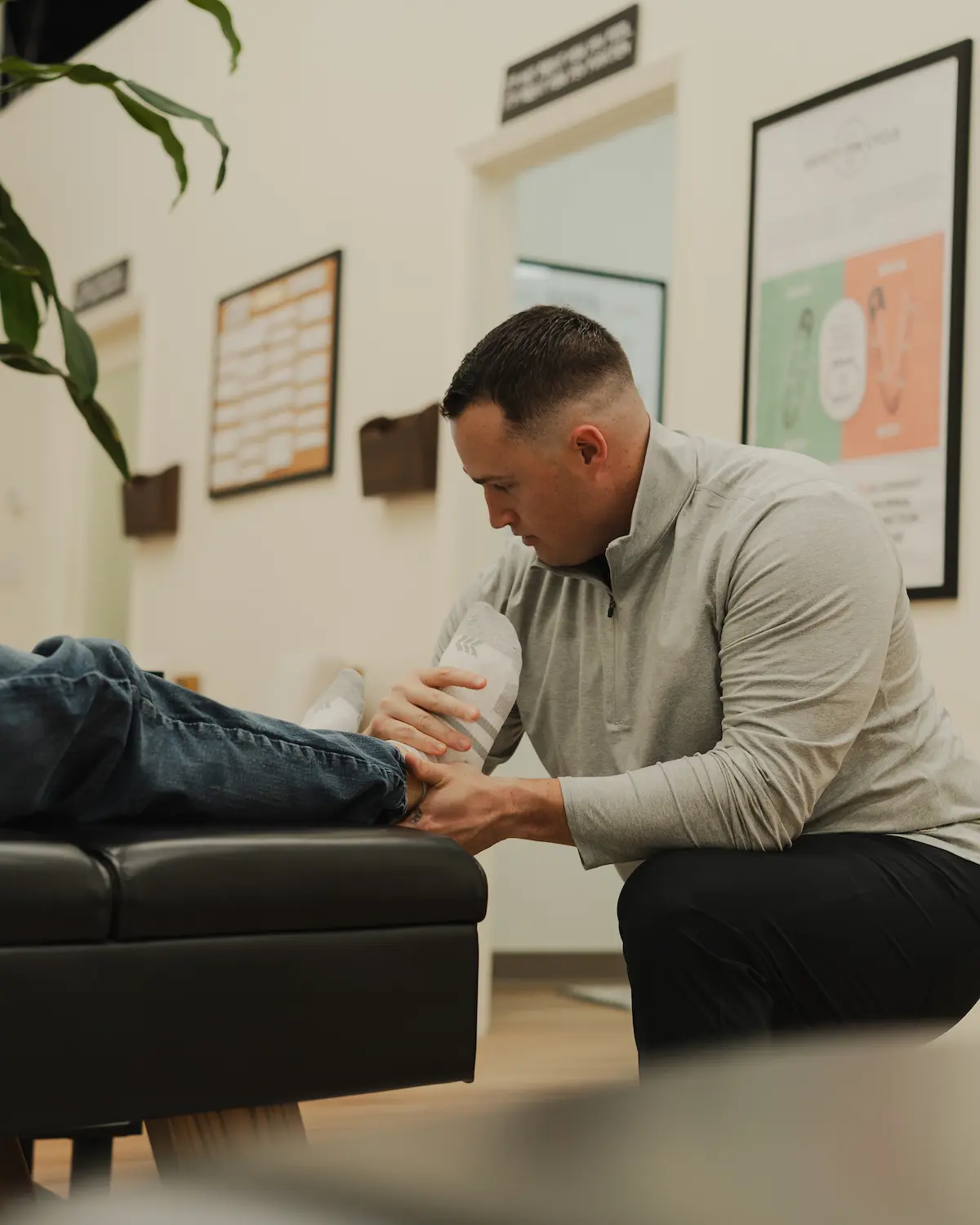 A male practitioner kneels, holding and examining a patient's foot on a black examination table in a clinic.