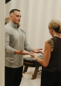 A man in a gray shirt and a woman with a tattoo on her shoulder face each other, gesturing with open hands in an indoor setting.