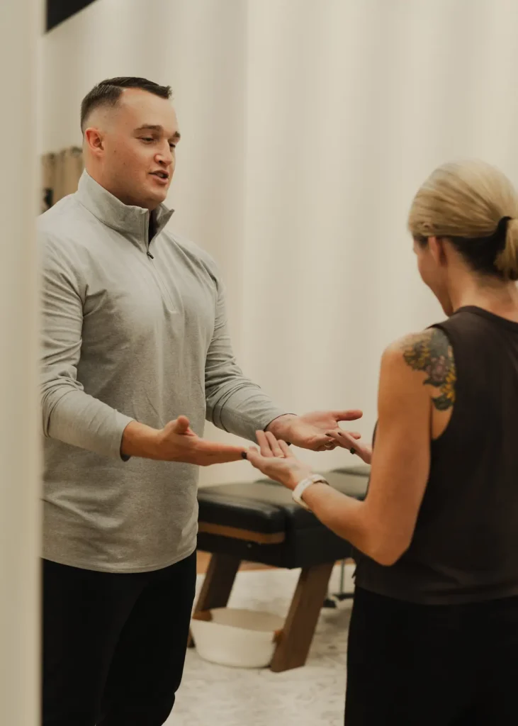 A man in a gray shirt and a woman with a tattoo on her shoulder face each other, gesturing with open hands in an indoor setting.