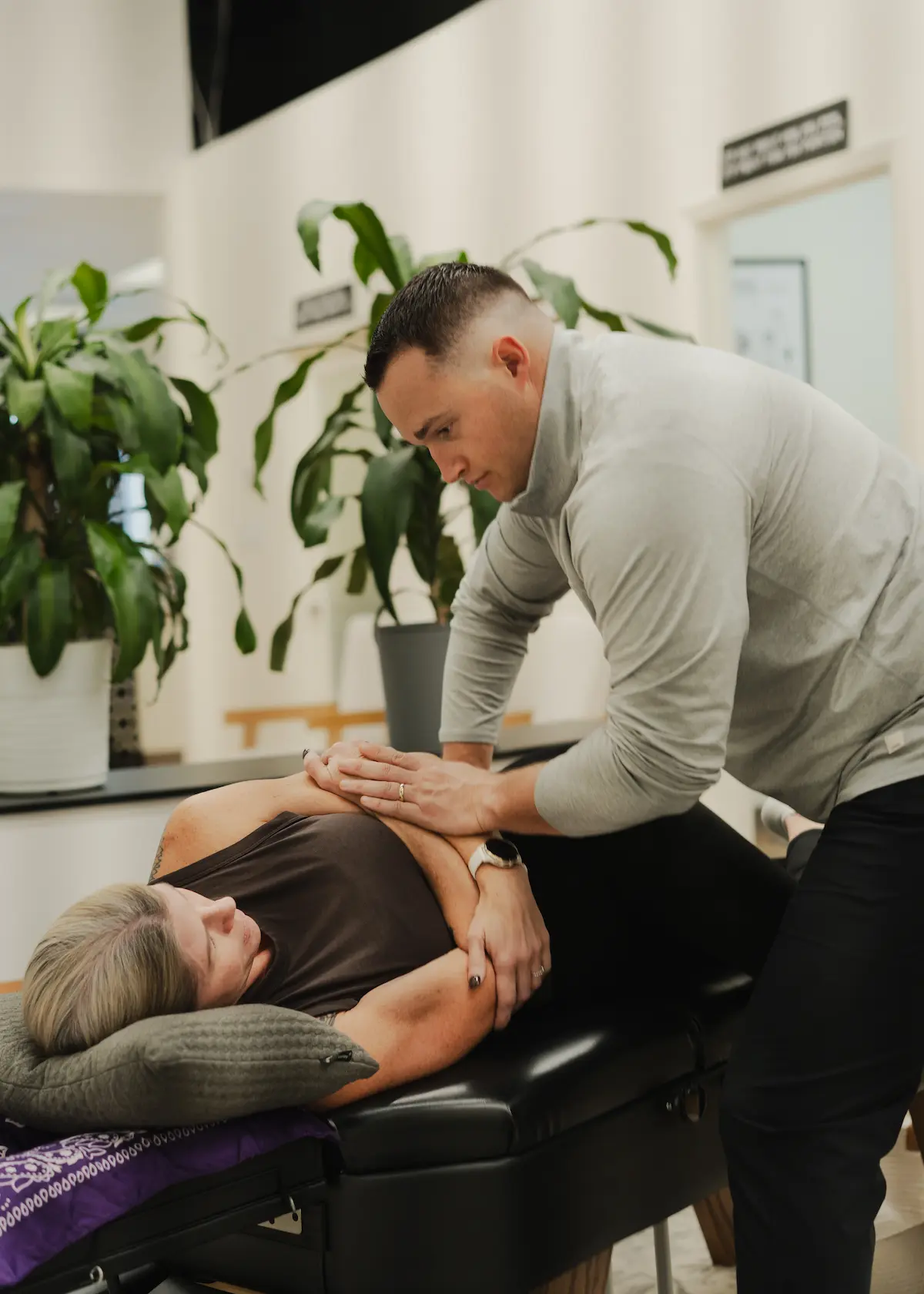A man in a grey shirt performs an adjustment on a woman lying on a black treatment table.