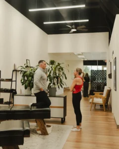 A man and a woman talk in a modern room with plants and wooden floors. The man leans on a black bench, and the woman stands with crossed arms.