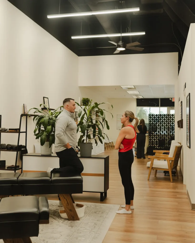 A man and a woman talk in a modern room with plants and wooden floors. The man leans on a black bench, and the woman stands with crossed arms.