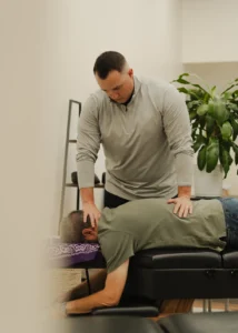 A male practitioner adjusts the neck of a male patient lying face down on a black examination table.