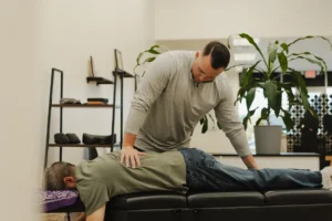 A male chiropractor adjusts the lower back of a male patient lying face down on a black treatment table.