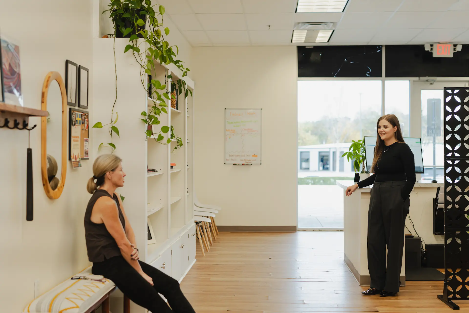 Two women in a bright office waiting area. One woman sits on a bench, and another stands behind a counter.