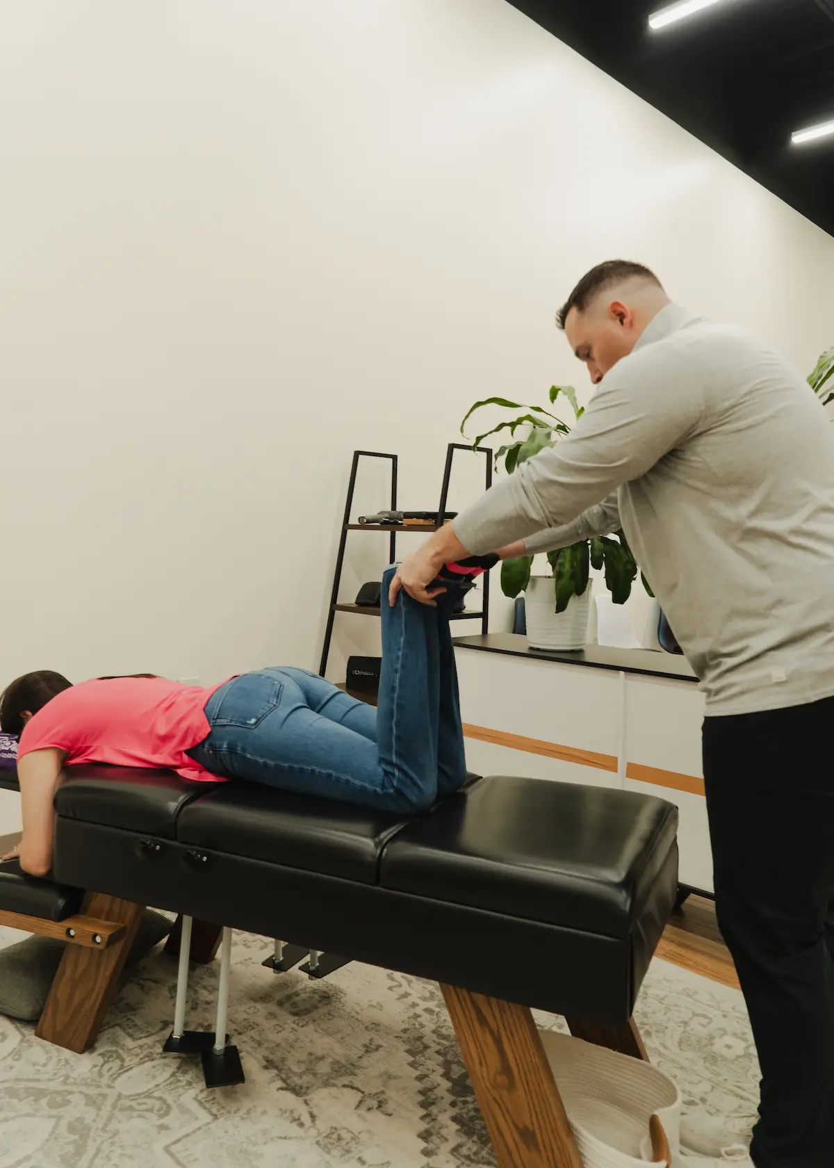 A male chiropractor adjusts the legs of a female patient lying face down on a black table.