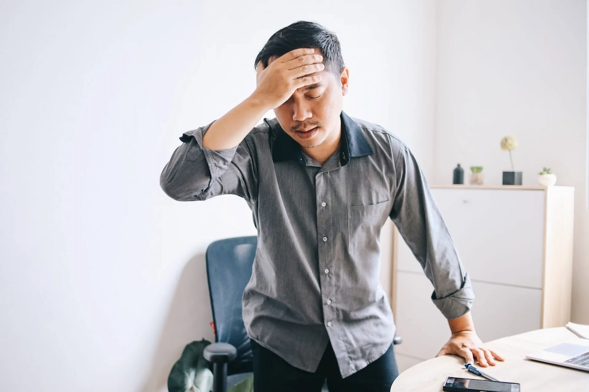 A man in a grey shirt stands with his hand on his forehead, looking distressed.