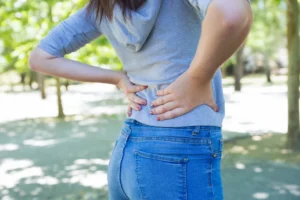 A person from behind, wearing jeans and a grey top, holding their lower back with both hands in an outdoor setting, indicating back pain.