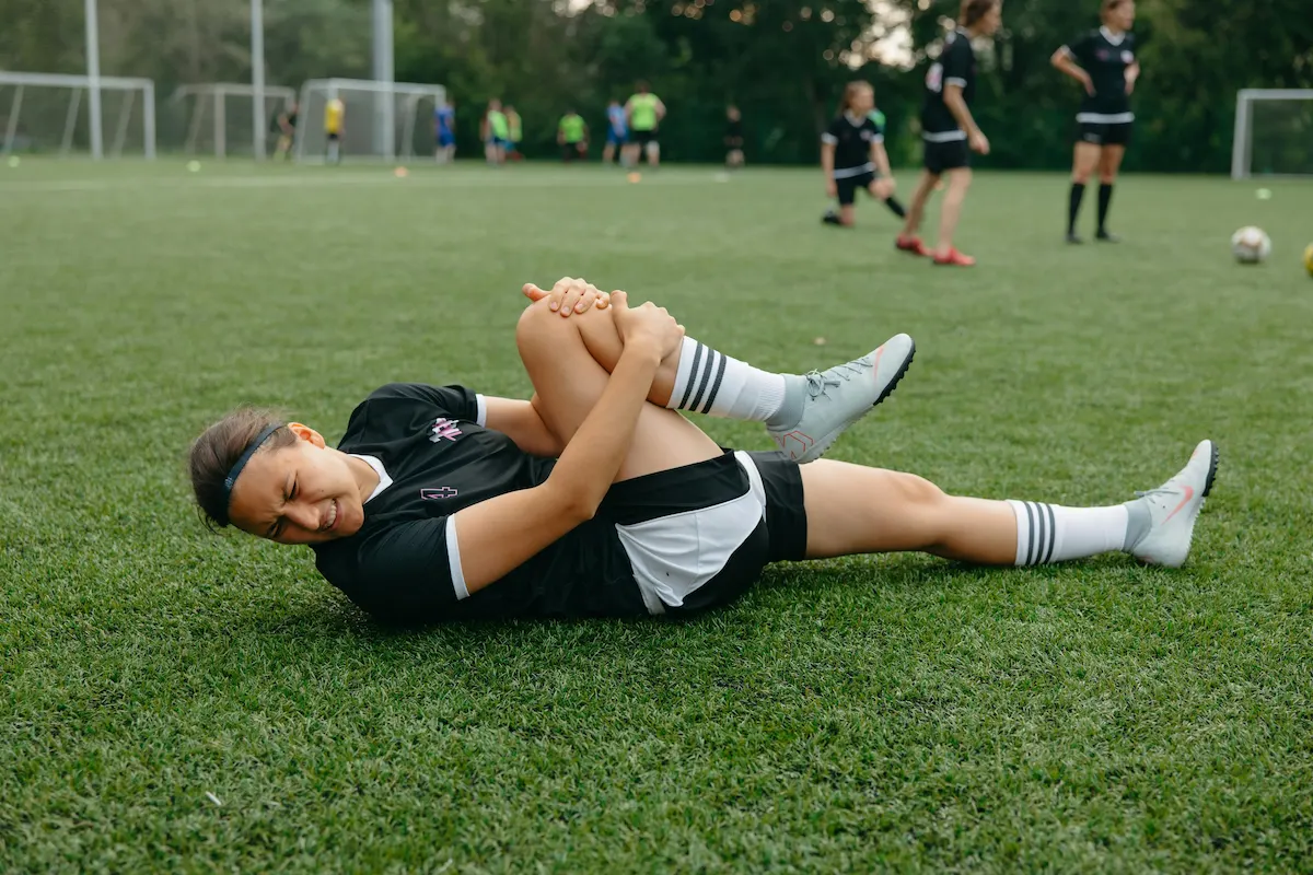A female soccer player lies on a green field, holding her knee with a pained expression.