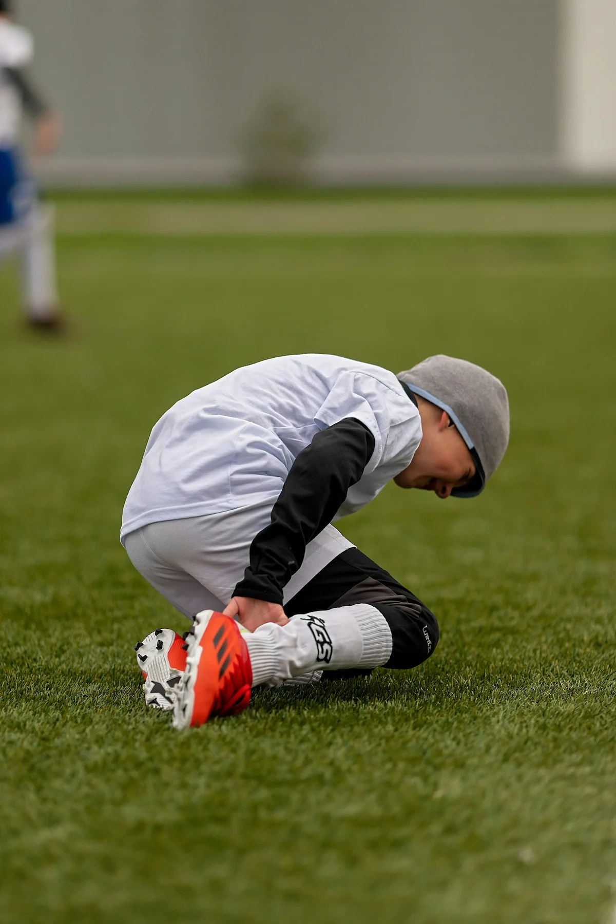 A young boy in soccer uniform crouches on a green field, holding his right ankle with both hands, appearing to be in pain.