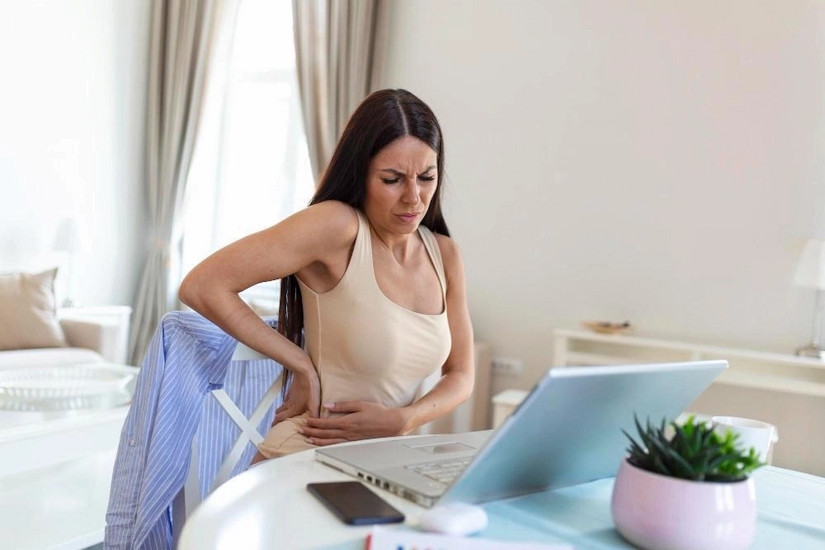 A woman in a beige tank top sits at a table, holding her lower back and stomach with a pained expression, next to a laptop.