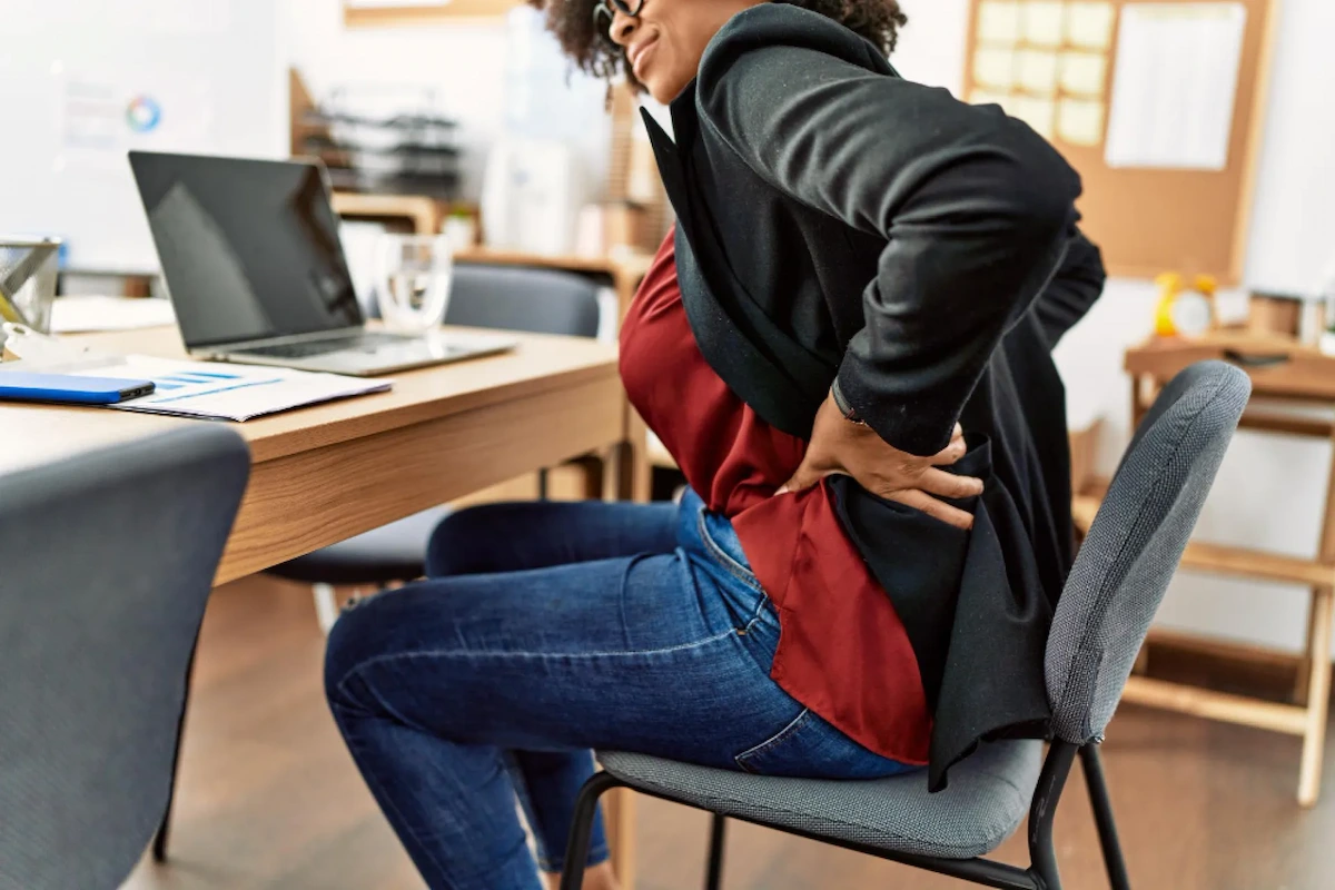 A person in an office chair holds their lower back in pain, with a laptop and documents on the desk.