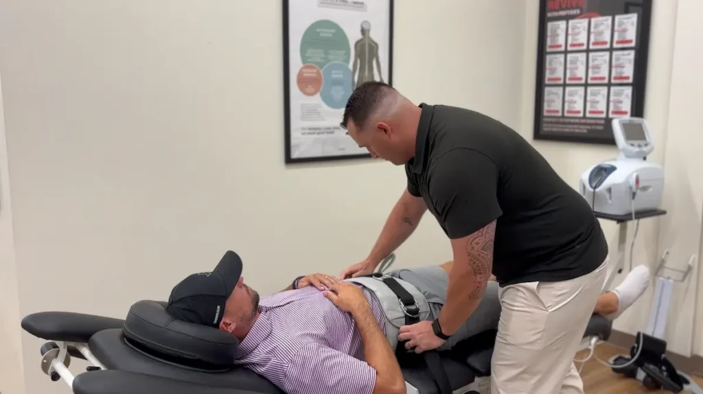 A practitioner in a black shirt adjusts a grey harness on a patient lying on a chiropractic table.