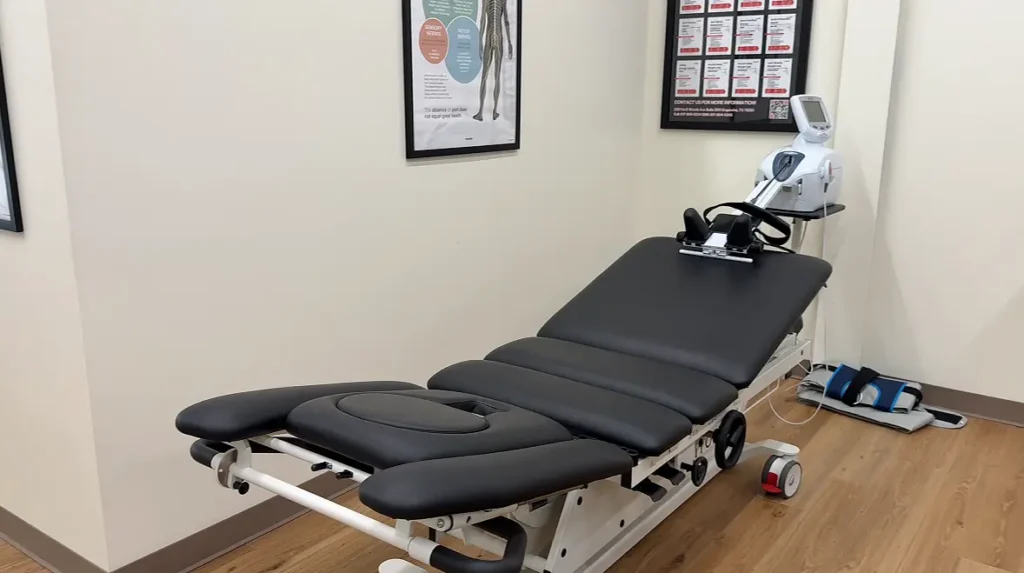 A black medical treatment table with a white and silver therapy device at its head, in a room with light walls and wooden floors.