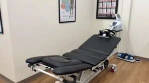 A black medical treatment table with a white and silver therapy device at its head, in a room with light walls and wooden floors.