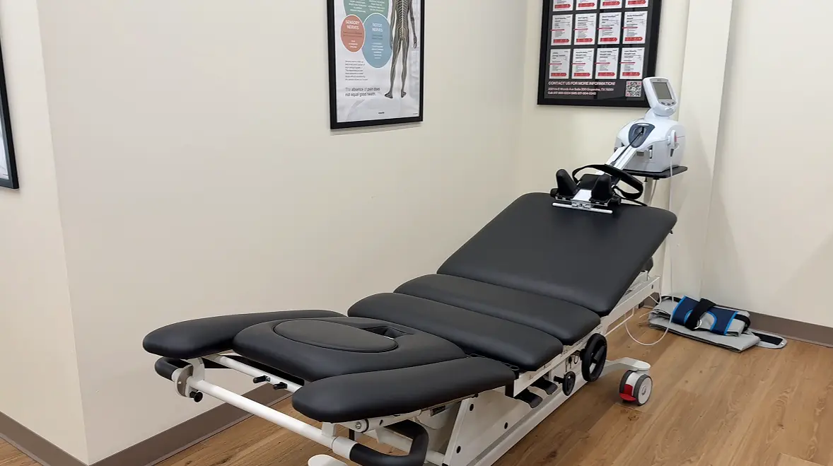 A black medical treatment table with a white and silver therapy device at its head, in a room with light walls and wooden floors.