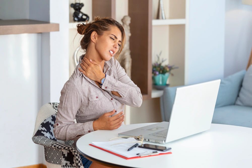 A woman with her hand on her neck and chest, showing discomfort while sitting at a table with a laptop.
