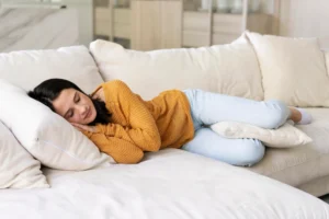 A young woman with dark hair sleeps soundly on her side on a light-colored sofa, wearing a yellow sweater and blue pants, with pillows supporting her head and legs.